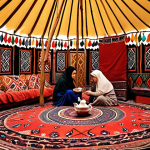 **Yurt Interior:** A warmly lit interior of a traditional Turkmen yurt (gara öý). Focus on intricate carpet patterns, colorful textiles, and family members sharing tea. Capture the sense of warmth, hospitality, and cultural heritage.